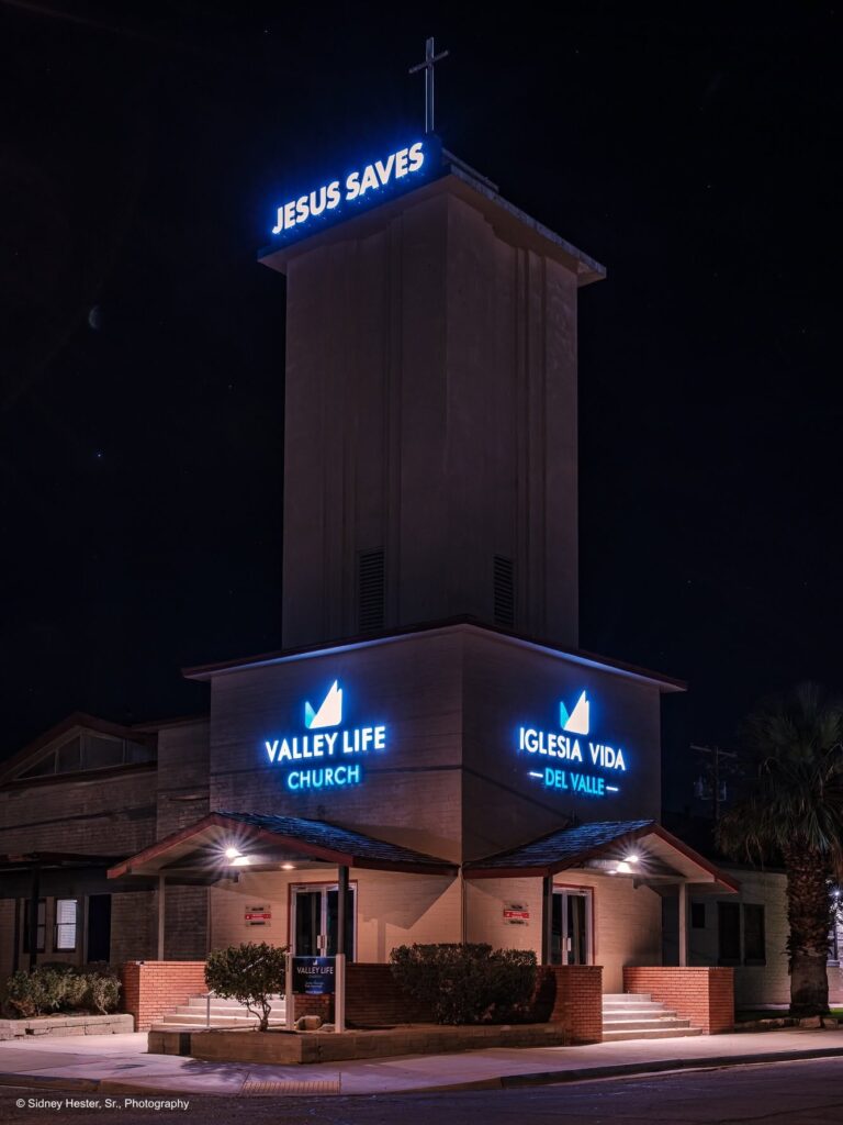 A nighttime photograph of Valley Life Church in El Centro, CA, with blue neon signs illuminated, including "JESUS SAVES" on the tower and "VALLEY LIFE CHURCH" and "IGLESIA VIDA DEL VALLE" over the entrances.